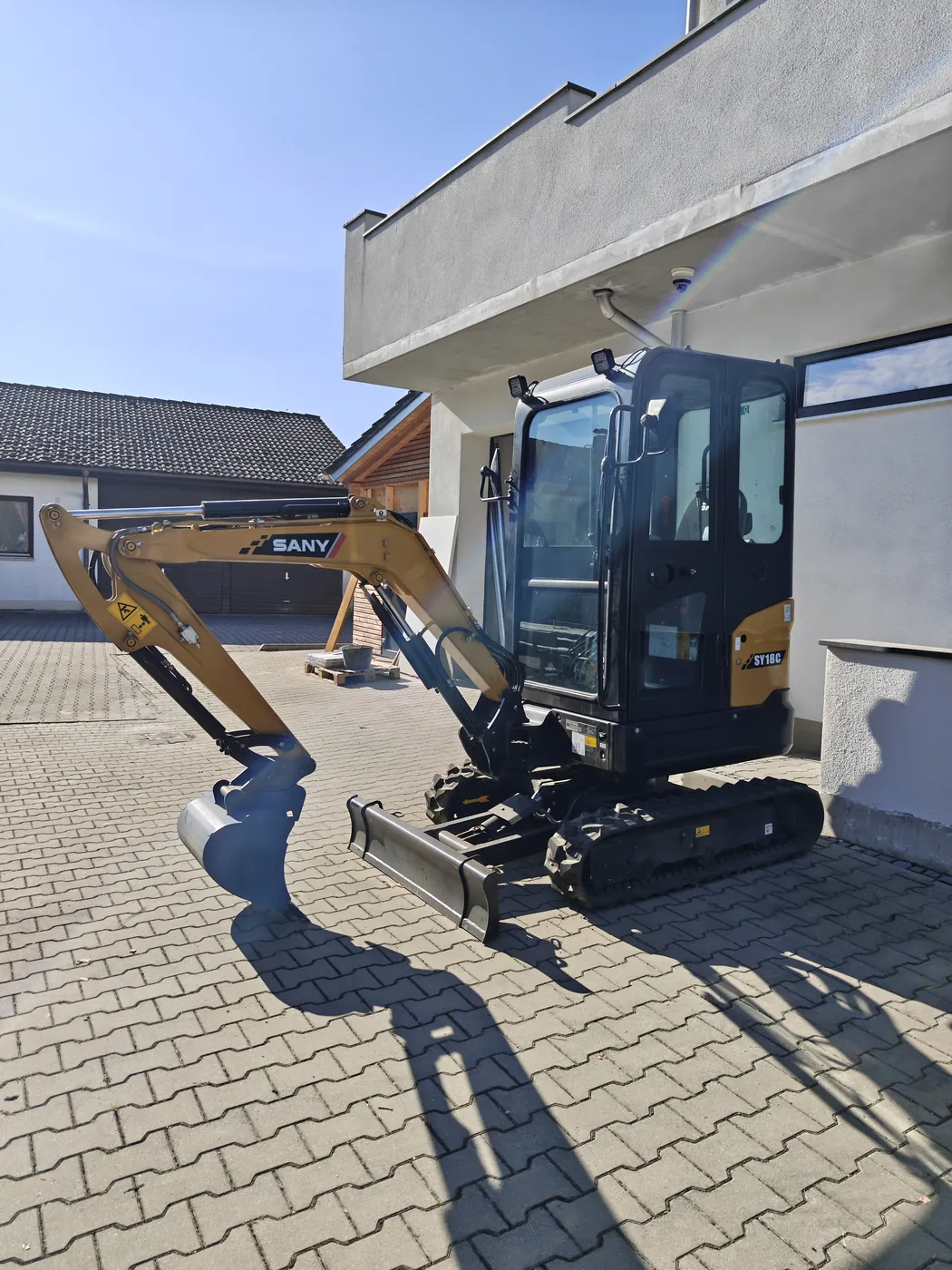 Minibagger SANY SY18C von W-RENT, schräg von vorn auf gepflastertem Hof: gelber Ausleger mit Tieflöffel, geschlossene Kabine, Planierschild und Gummiketten.