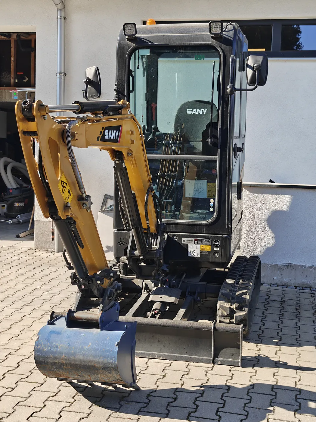 Minibagger SANY SY18C von vorn: Tieflöffel am Boden, Planierschild quer davor, LED-Arbeitsscheinwerfer und Rundumkennleuchte auf dem Kabinendach.